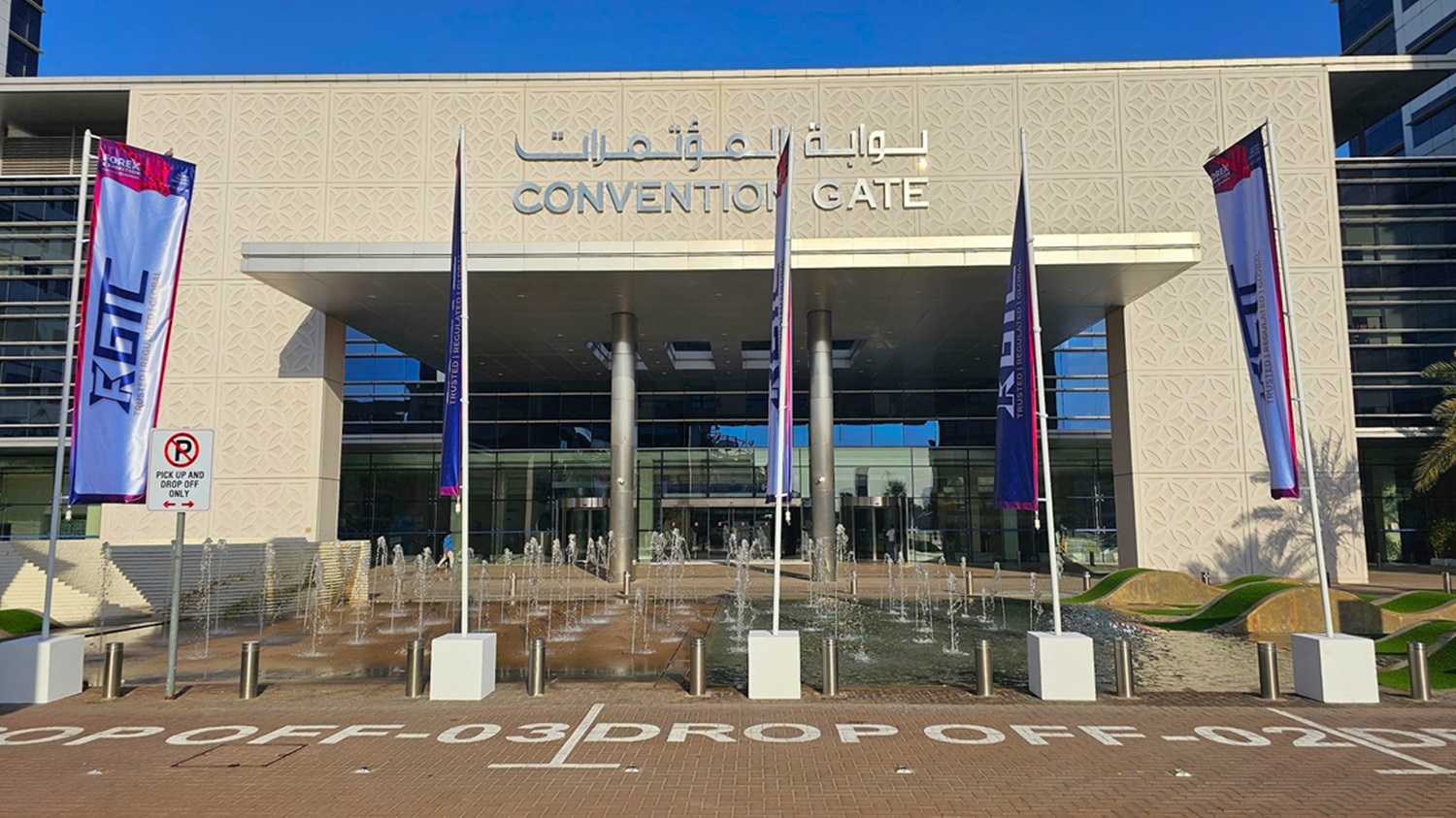 A modern building with a glass facade on the right, featuring large windows and a sleek design. On the left, there are palm trees lining a paved walkway. The building has a decorative patterned wall and a large overhang supported by metal columns. The ground is covered with interlocking bricks, and there are bollards along the path. Shadows of the palm trees are cast on the ground.