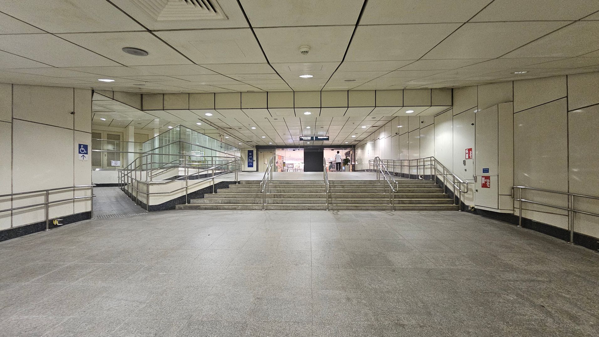 A spacious underground passage with a grey tiled floor and a light-coloured panelled ceiling. In the centre, a set of stairs with metal railings leads upwards, flanked by ramps on both sides for accessibility. The walls are lined with light panels, and there are signs indicating directions. At the top of the stairs, a glass entrance is visible, with a person standing near it. A fire extinguisher is mounted on the right wall.