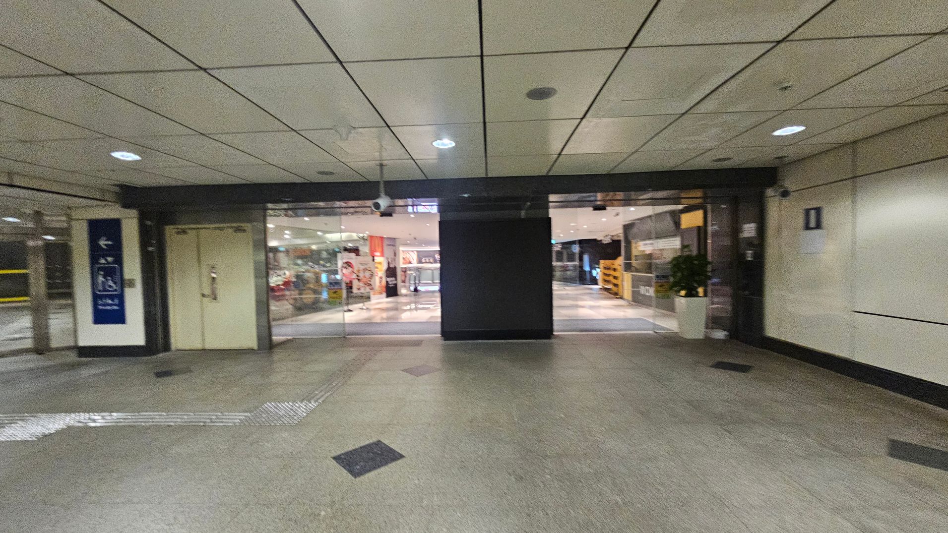 A corridor with a tiled ceiling and greenish floor. In the centre, there are glass doors leading to a brightly lit interior space with visible signage and displays. On the left, there's a sign with directional arrows and a closed door. On the right, a wall with a small sign and a potted plant near the entrance. The area is well-lit with ceiling lights.