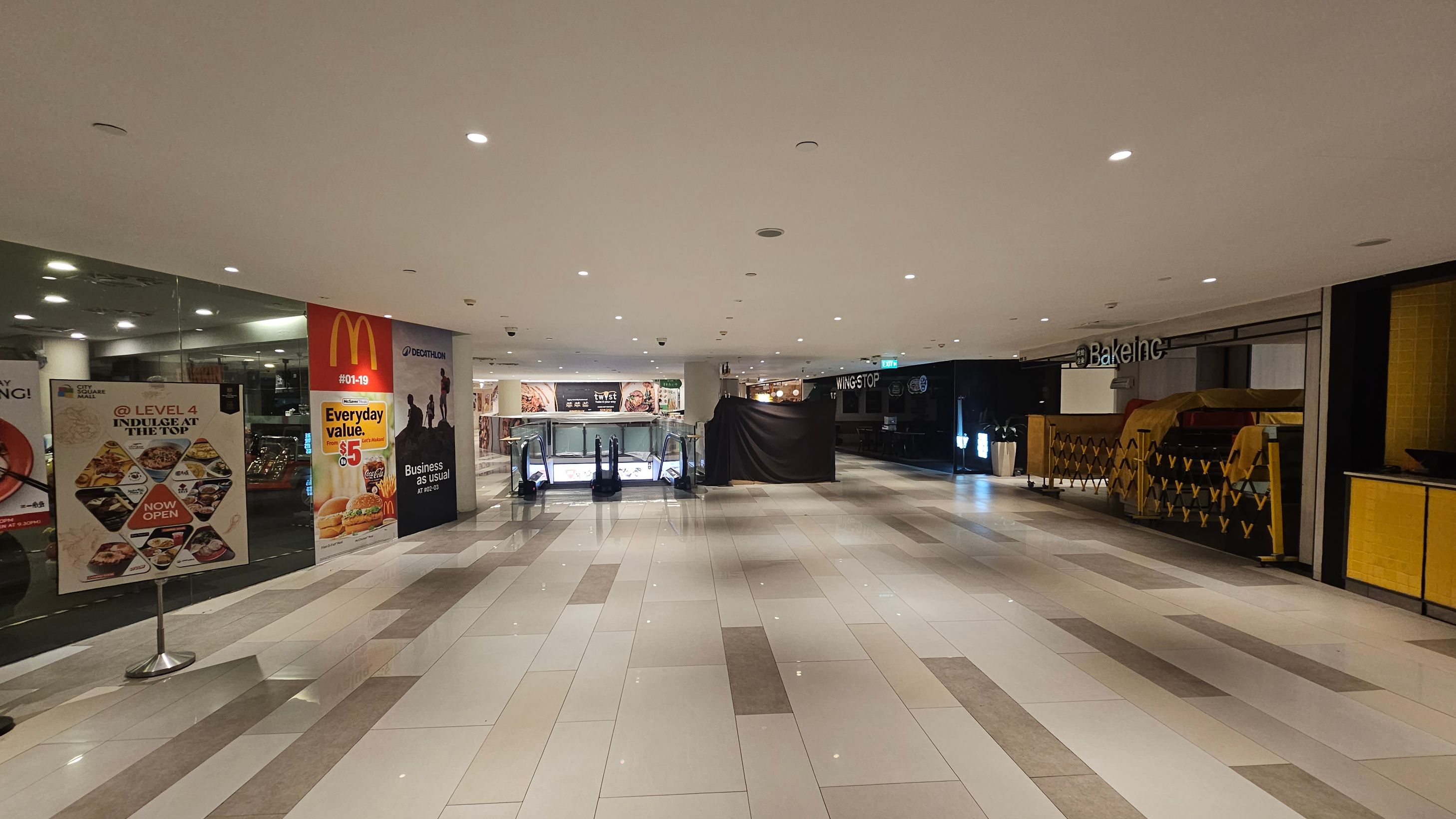 A shopping mall interior with a wide, tiled floor in shades of grey and white. On the left, there are promotional posters, including one for McDonald's. In the centre, an escalator is visible, leading to a lower level. On the right, there is a closed shop with a sign reading "Bake Inc" and a covered counter. The ceiling is white with recessed lighting, creating a bright atmosphere.