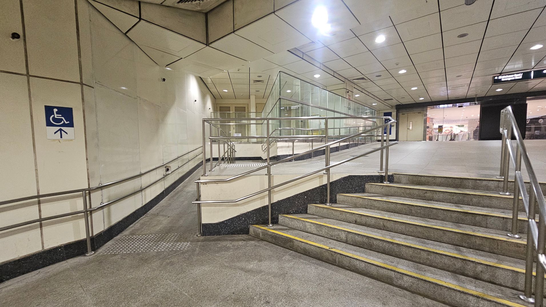 A public indoor space with a ramp and stairs. On the left, a ramp with metal railings leads upwards, marked by a blue sign indicating accessibility. The right side features a set of stairs with similar metal railings. The background shows a glass partition and a view into a well-lit area with shops. The ceiling is tiled with recessed lighting, and the floor is a mix of green and grey tiles.