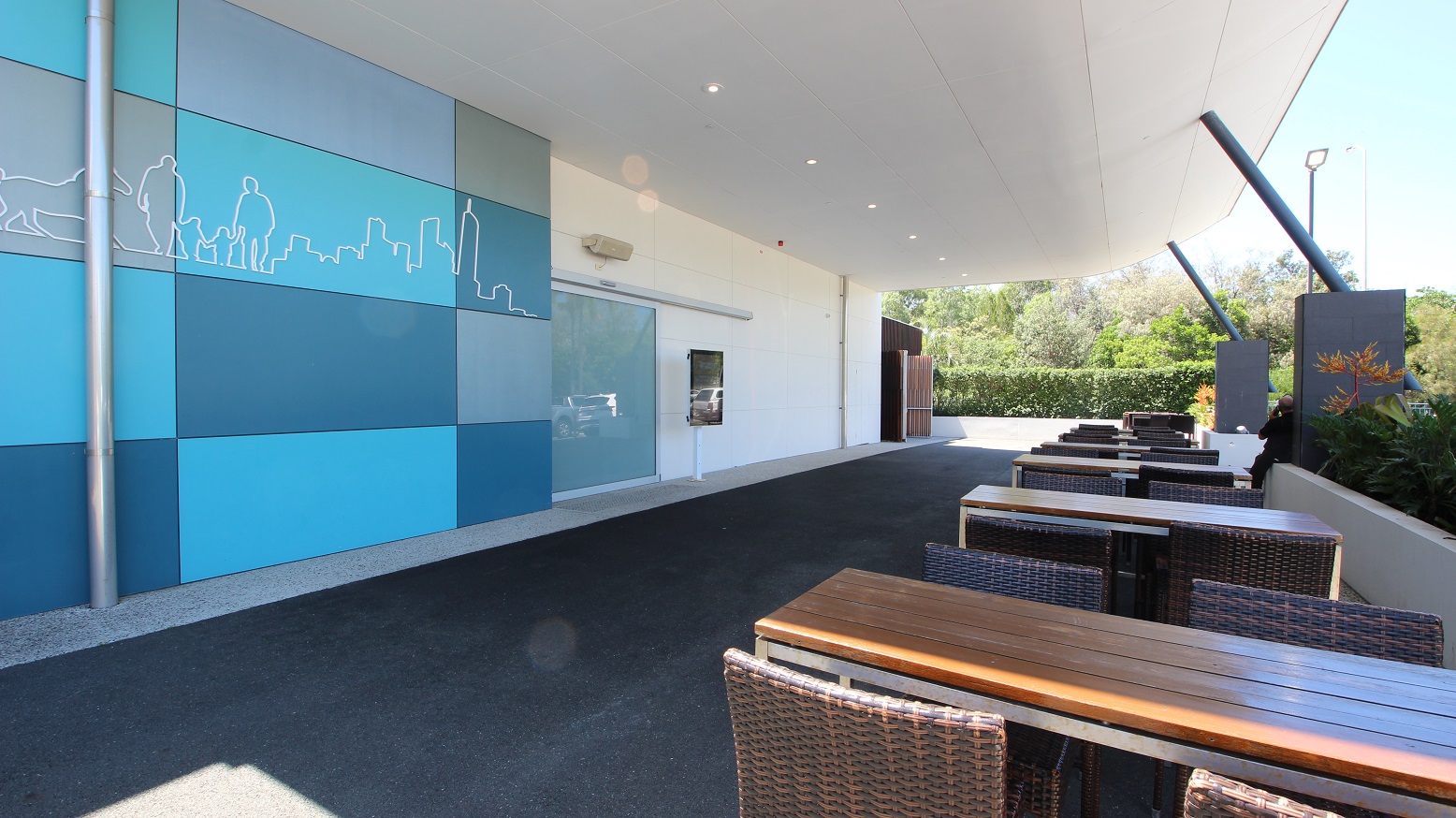 Drop off point for the Sea World Resort Conference Centre showing the 3 accessible car parks on the left hand side of the facility and many regular carparks directly in front of the building.  This image is from a side angle of the building.  The conference centre is a single story building with large grey and blue tiled exterior.  A large portico offers under cover drop off areas.