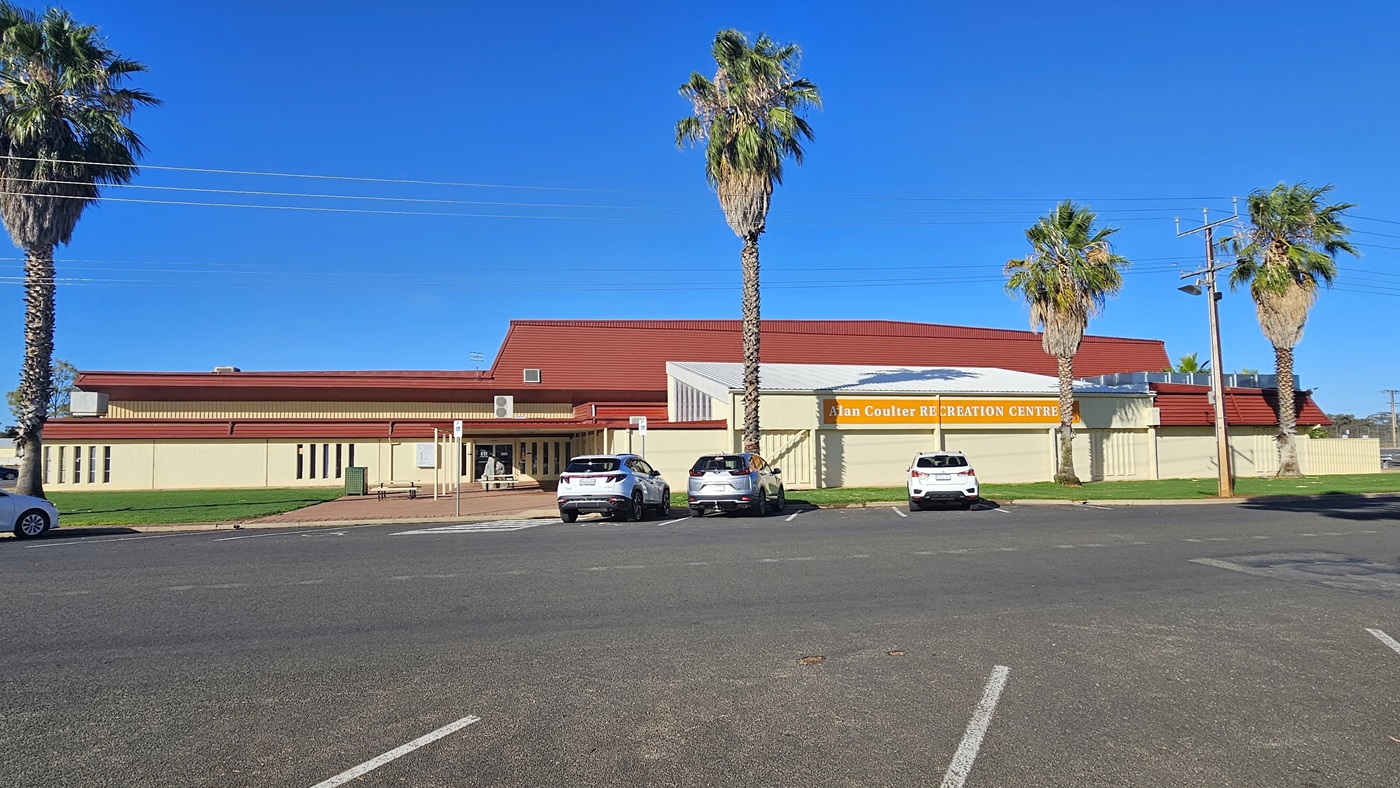 A cream industrial building with red corrugated roofing. In front is a parking area with four cars of various colors and models parked. In the middle, the facility's signage in yellow and white bold letters is visible. Tall palm trees border the parking area. The building's concourse has red bricks and manicured grass.