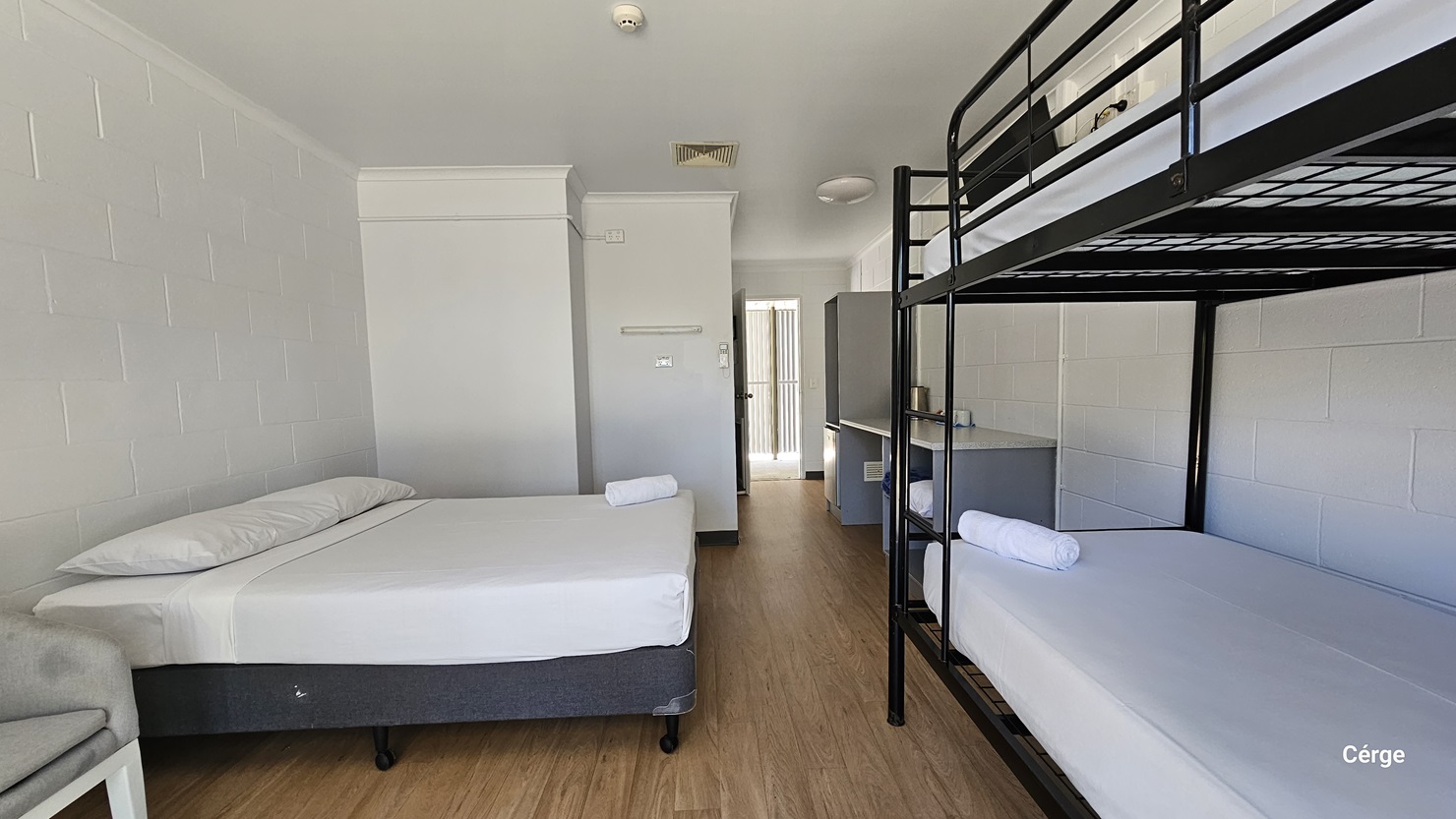 Inside the room, to the left is a double bed with white linens. Beside it is a cushioned grey chair. To the right is a double-deck bed. Adjacent to it is a table holding an electric kettle and cups. The ceiling and walls are all white with a brick design. The floor has a timber design.
