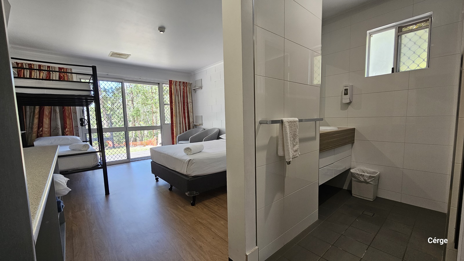 Inside the room, to the left is a double bed with white linens. Beside it is a cushioned grey chair positioned near the glass window. To the right is a double-deck bed. Adjacent to it is a table. The ceiling and walls are all white with a brick design. The floor has a timber design. To the right is the ensuite toilet featuring grey tiles and white walls. A washbasin and rubbish bin are positioned in the corner.