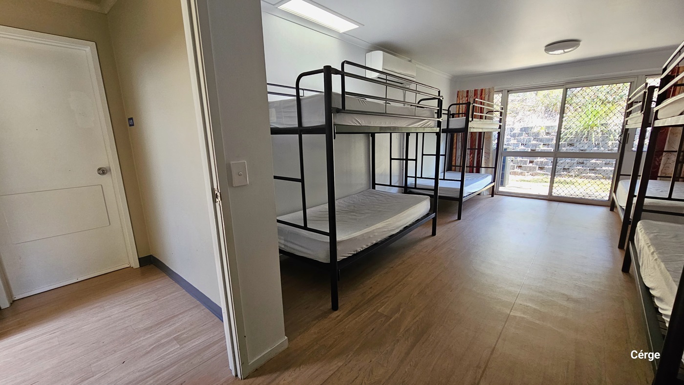 To the left is a white door. To the right, there are four double-decker beds with white linens. The wall on the right is made of glass and covered with floor-to-ceiling curtains. The ceiling and walls are white with fluorescent lights. The floor has polished timber.