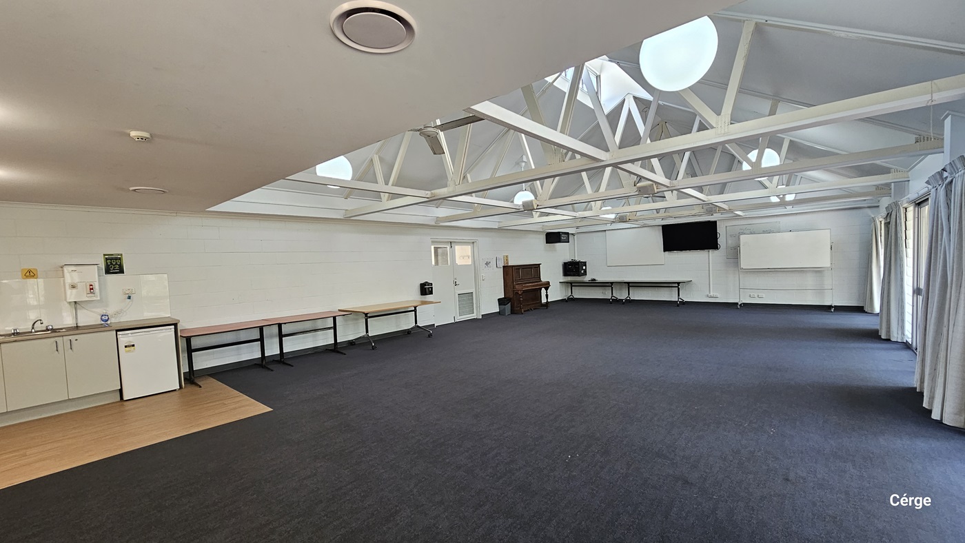 Inside the meeting room, a drinking fountain is situated to the left. A small section of timber tiles is installed on the floor. Tables are arranged against the wall. At the far end, there is a wooden piano, a flatscreen TV, and a movable whiteboard. To the right, floor-to-ceiling curtains cover the glass wall. The ceiling features an industrial look with exposed white metal beams and pendant lights. The floor has dark grey tiles.