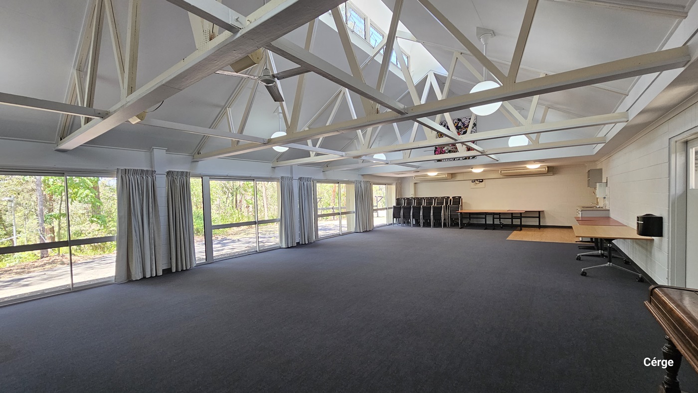 Inside the meeting room, a drinking fountain is situated to the far right. Tables and plastic chairs are arranged against the wall. To the left, floor-to-ceiling curtains cover the glass wall. The ceiling features an industrial look with exposed white metal beams and pendant lights. The floor has dark grey tiles.