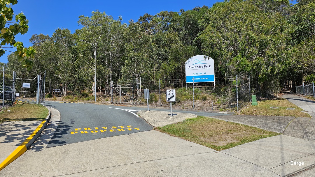 The concrete road splits, with one path leading right towards a gated fence. On the left, a larger road is marked "Private Property" in yellow. A prominent white and blue sign displays the facility's name to the right. Trees line the road and surround the area. A chain fence and gate secure the perimeter, while road signs and tall street lights are visible.