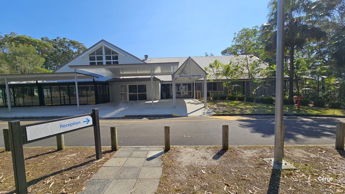 A single-storey building with a gabled roof is in the centre, featuring large windows and a covered entrance. To the left, there is a reception sign with an arrow pointing right. On the right, a tall palm tree stands near a lamppost. The foreground has a paved walkway and wooden posts, while the background includes trees and greenery. The floor is paved with concrete tiles, and the ceiling of the entrance area is white and flat.