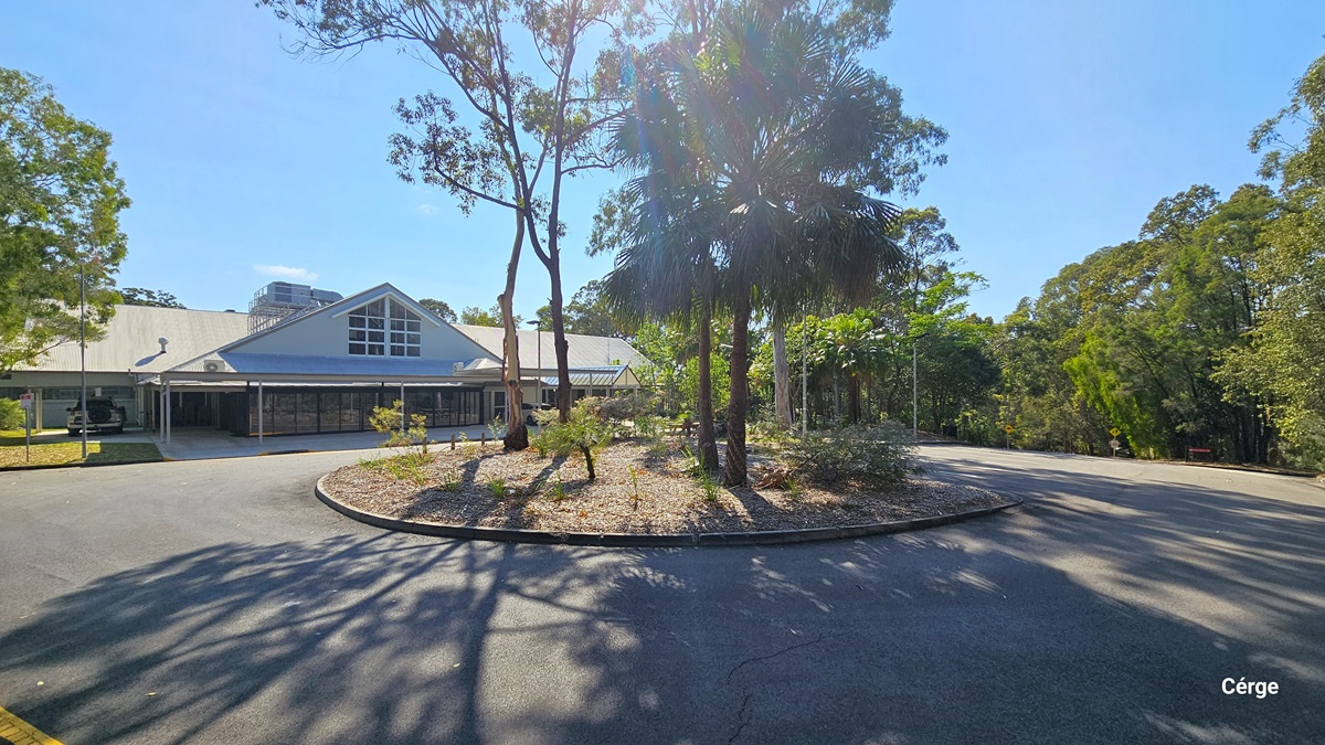 A paved area with a circular garden bed in the centre, surrounded by a road. Sparse trees and shrubbery populate the garden bed and the periphery. Behind is a building with a large triangular roof feature and an extended wing. Tall green trees frame the background.