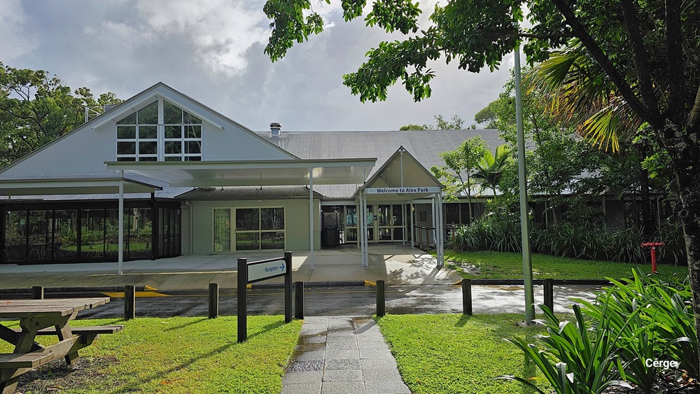 A building with a gabled roof and large windows on the left, featuring a covered entrance with a sign reading "Welcome to the Park" in the middle. The right side has lush greenery, including trees and bushes. In the foreground, there is a picnic table on a grassy area and a paved walkway leading to the entrance. The background includes more trees and foliage. The floor is a mix of grass and pavement.