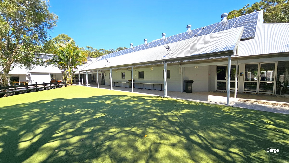 An exterior view of a modern single-storey building with floor-to-ceiling windows and doors. An overhanging roof shades a concrete walkway extending towards the foreground. To the right, a landscaped area features trees and bushes. Behind is a fenced area with synthetic turf.