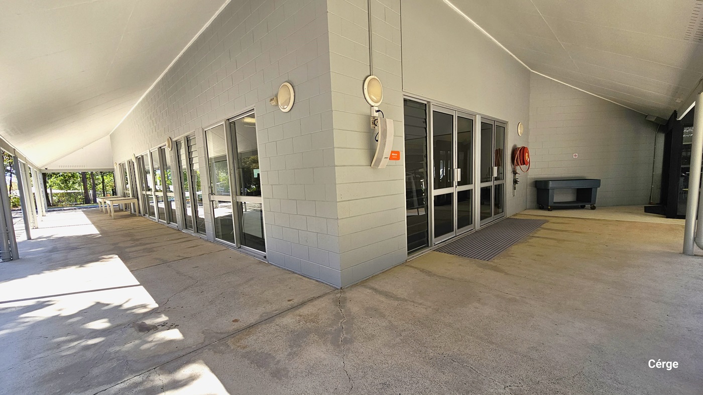 A wide-angle view of a modern building's entrance, featuring a covered walkway with concrete floor tiles leading up to a set of double glass doors. The building has light grey exterior tiles and multiple rectangular windows. To the right of the doors, a red fire hose reel is mounted on the wall, and a black trash bin stands nearby. Light fixtures are attached to the wall.
