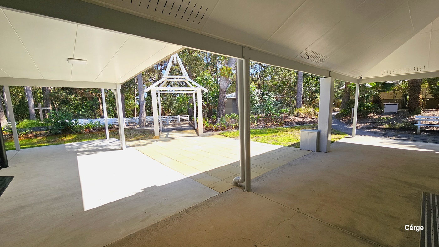 An outdoor covered patio area featuring a large open space with a concrete floor, leading to a garden with lush greenery. In the centre, there is a white wooden arbour, while seating areas and a barbecue station are visible in the background under the shade of trees.