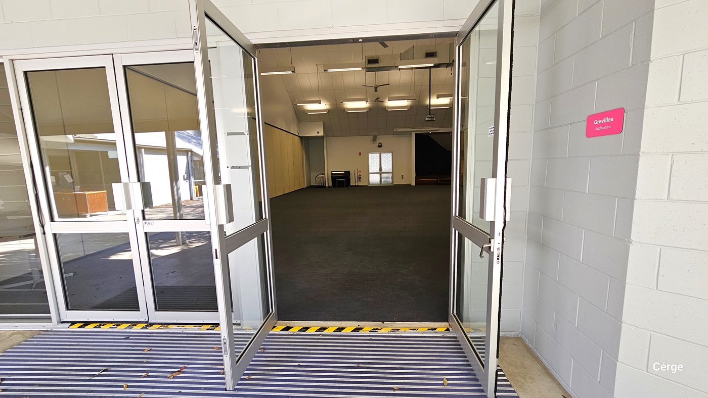 A building entrance with a set of double glass doors that opens outward leading into a spacious interior with dark carpeting. The ceiling has fluorescent lights and A non-slippery mat is positioned before the door. To the right of the doors, affixed to the grey brick wall, is a pink sign with the word 'Grevillea Auditorium' written in white letters. The threshold features yellow and black hazard warning stripes, indicating caution for stepping or wheeling over.