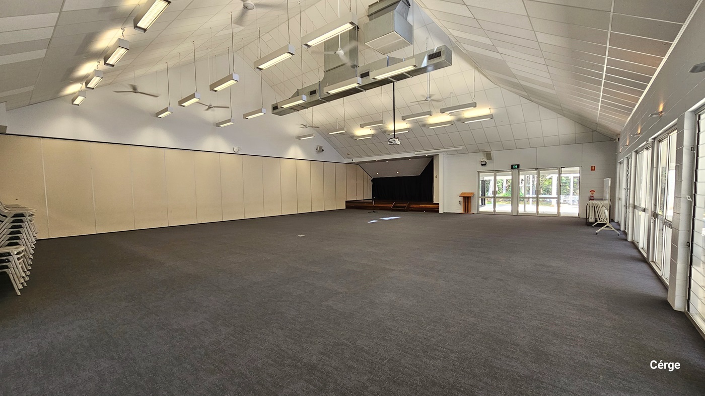 A spacious, empty hall with grey carpet, cream partitions, a pitched ceiling with fluorescent lights, and ceiling fans. A small raised stage is at the far left. A door is visible on the right, and adjacent to it is a fire extinguisher and wooden table.