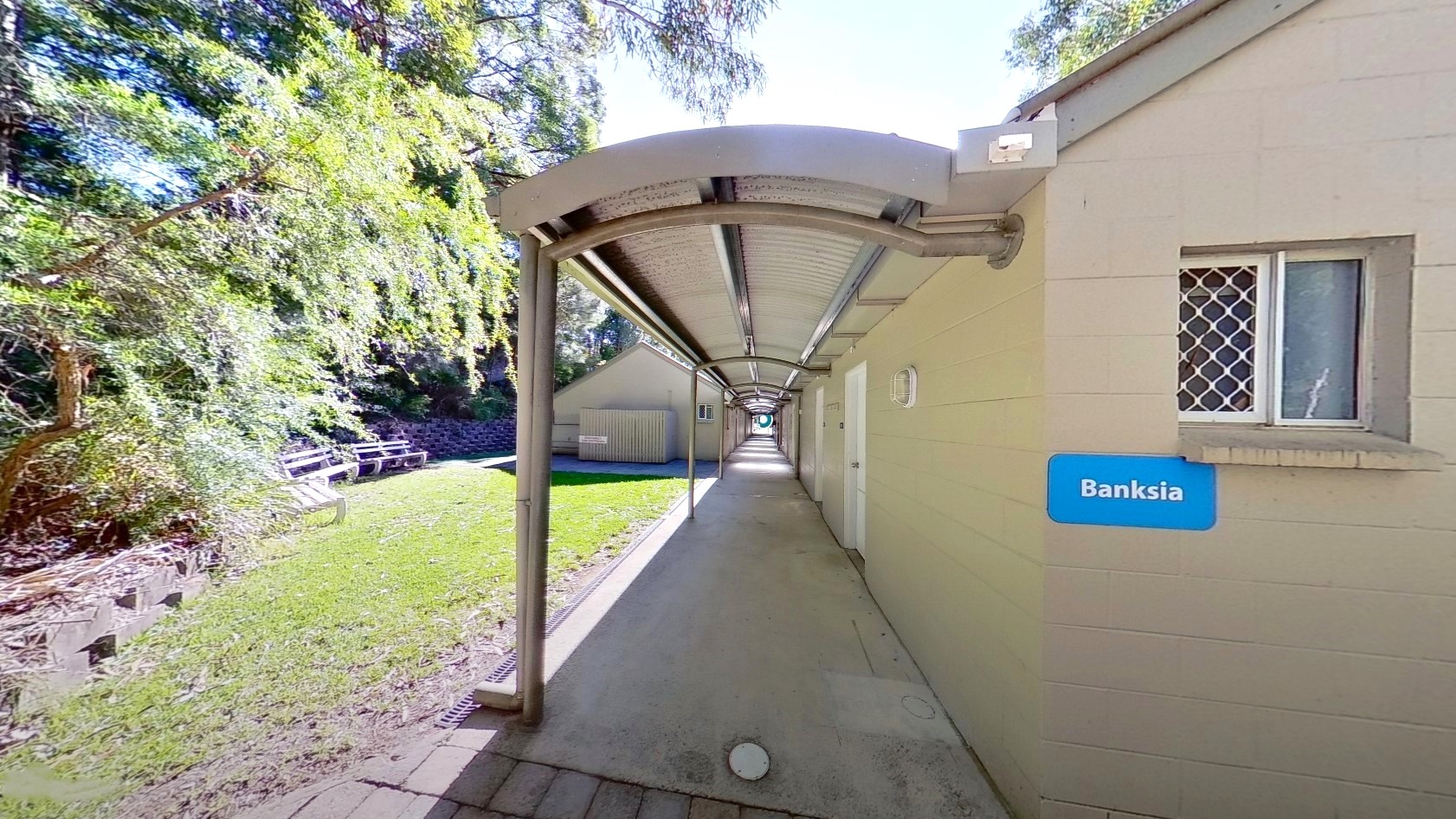 The Banksia accommodation is a low-rise cream building with a small window and an extended roofing. To the left, trees and grass surround the area. Bench seatings are arranged under the shades of huge trees.