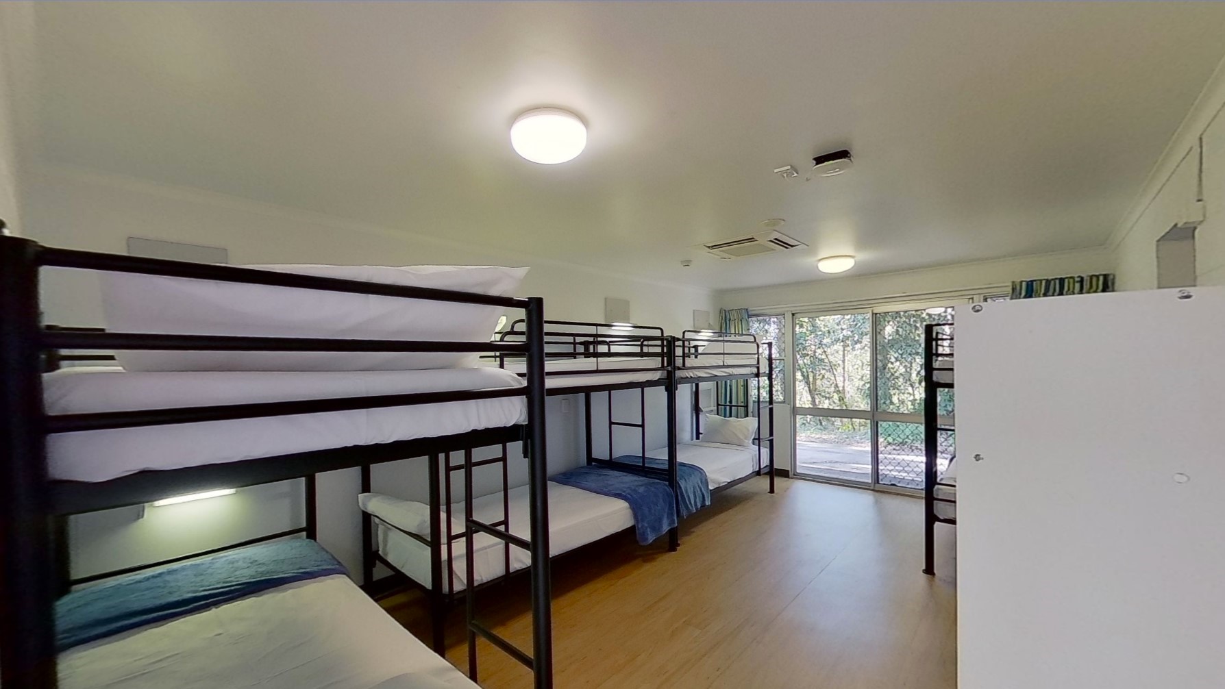 Bunk beds with white and blue linens are positioned on the left, with another set near the wall on the right. Floor-to-ceiling curtains are affixed to the glass wall. The floor is made of timber, and the ceiling and walls are all white.