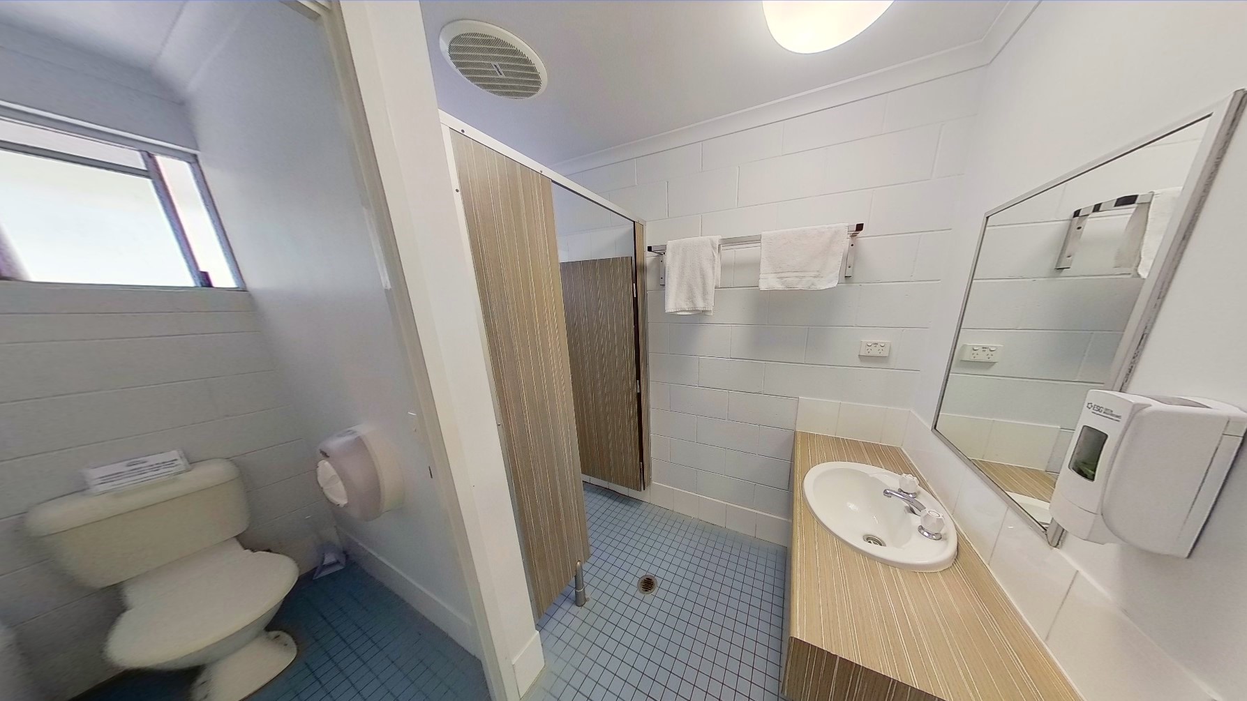 To the right is a toilet bowl with a tissue dispenser. Beside it is a lavatory with a wooden design. A liquid soap dispenser and mirror are affixed to the wall. A door with a wooden design secures a bathroom stall. A stainless bar holds two white towels. The walls and ceiling are all white with LED lighting. The floor has powder blue tiles.