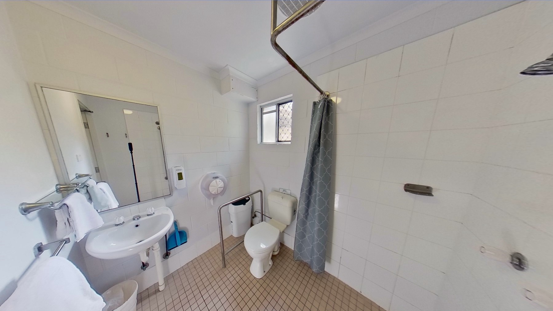 To the right is a wash basin, with mirrors and a stainless steel bar holding white towels. Underneath is a rubbish bin. In the corner is a toilet bowl equipped with hand grabs and another rubbish bin. A tissue dispenser and soap dispenser are mounted to the wall. A curtain is suspended from a bar, securing the shower area. The walls and ceiling are all white, and the floor has cream tiles.
