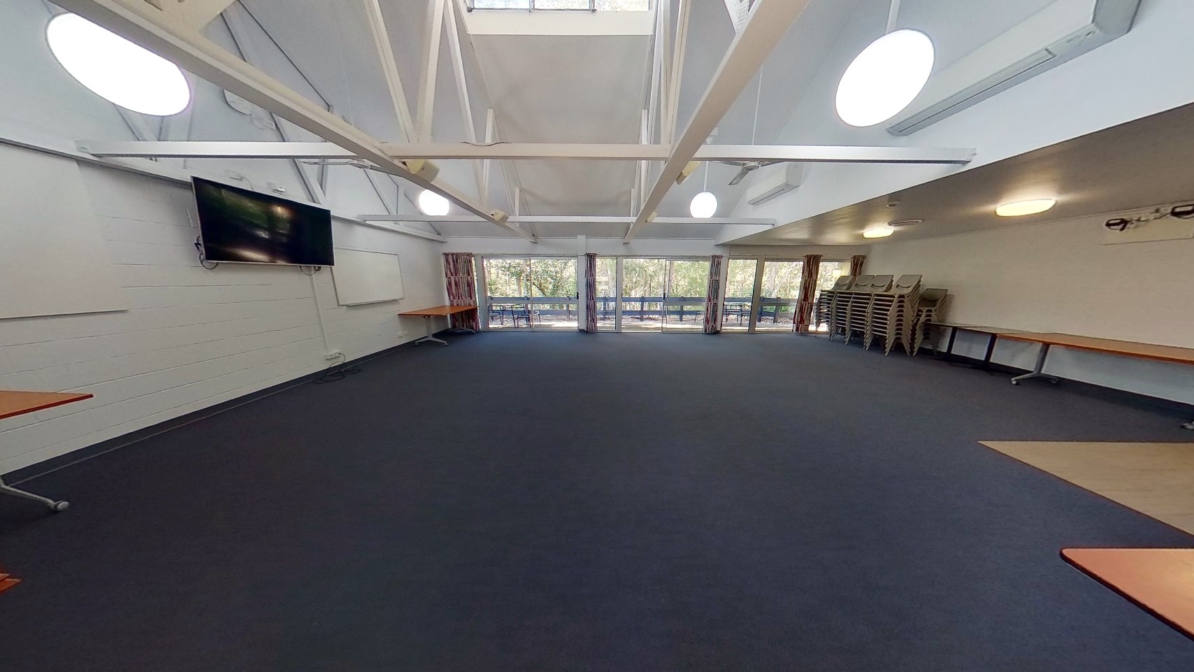 Inside the meeting room, tables and plastic chairs line the right wall. A flatscreen TV and a whiteboard are mounted on the left wall. The ceiling boasts an industrial look with exposed white metal beams and pendant lights. Dark grey tiles cover the floor. At the far end is a floor-to-ceiling glass window with curtains.