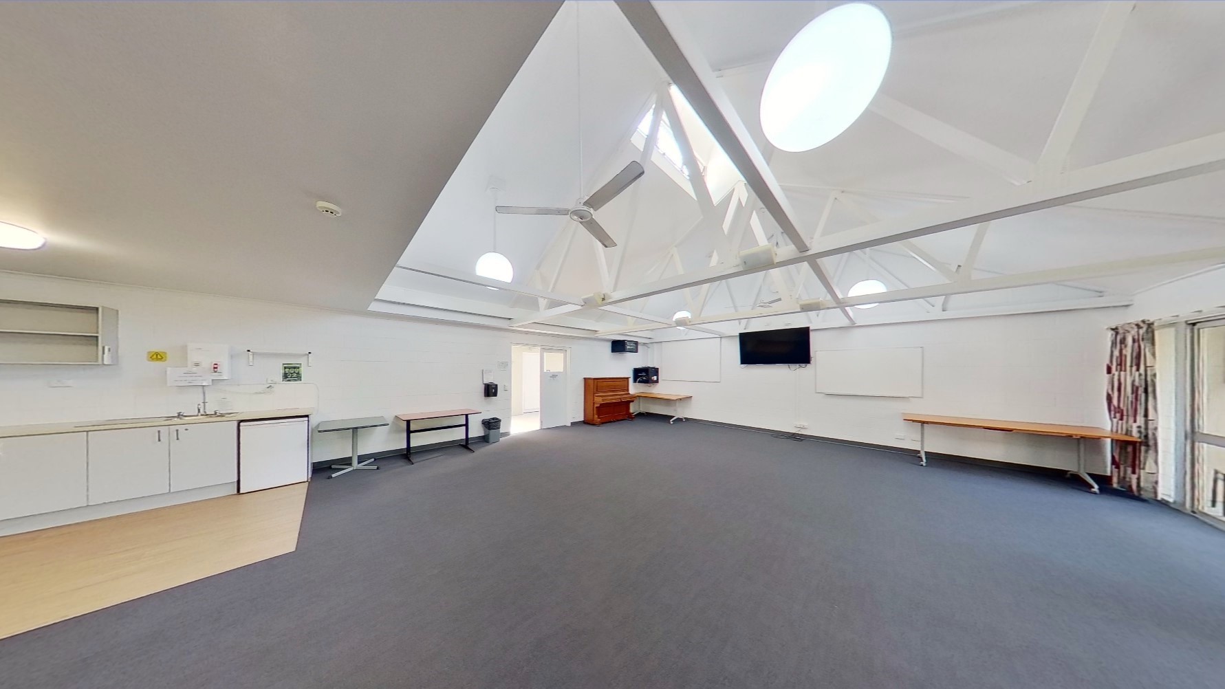 Inside the meeting room, a drinking fountain is positioned on the left. Tables line the left wall, with a flat-screen TV and a whiteboard mounted on the middle wall. A wooden piano sits in the corner. At the far end is a floor-to-ceiling glass window with curtains. Additional tables are against the right wall. The ceiling boasts an industrial look with exposed white metal beams and pendant lights. Dark grey tiles cover the floor.