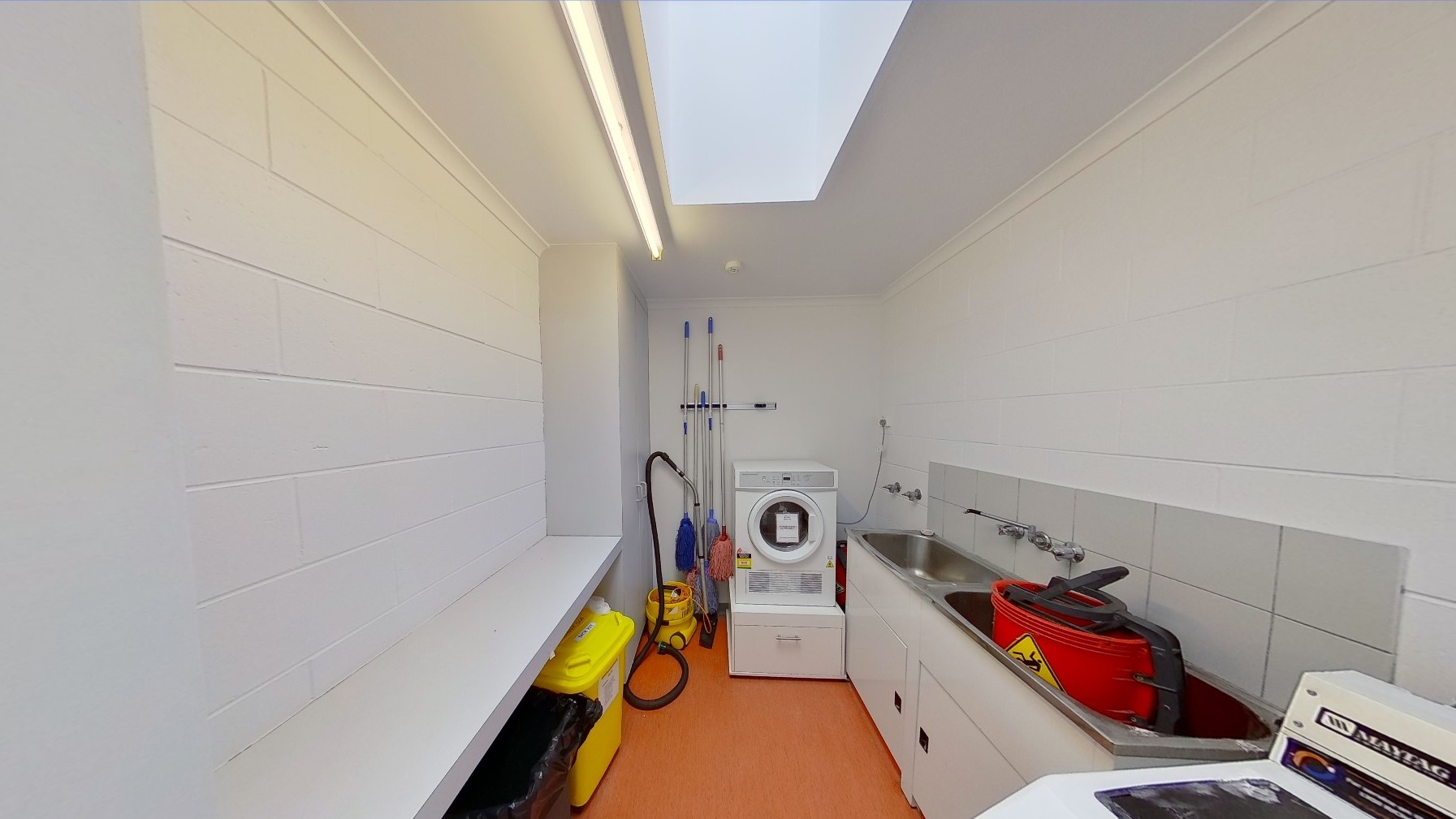 To the left, cleaning materials and rubbish bins are placed under a wooden ledge. To the right is a washing machine and a lavatory with a cleaning bucket. In the middle is another washing machine and other cleaning materials. The wall and ceiling are all white with a huge fluorescent light, and the floor is made of timber.
