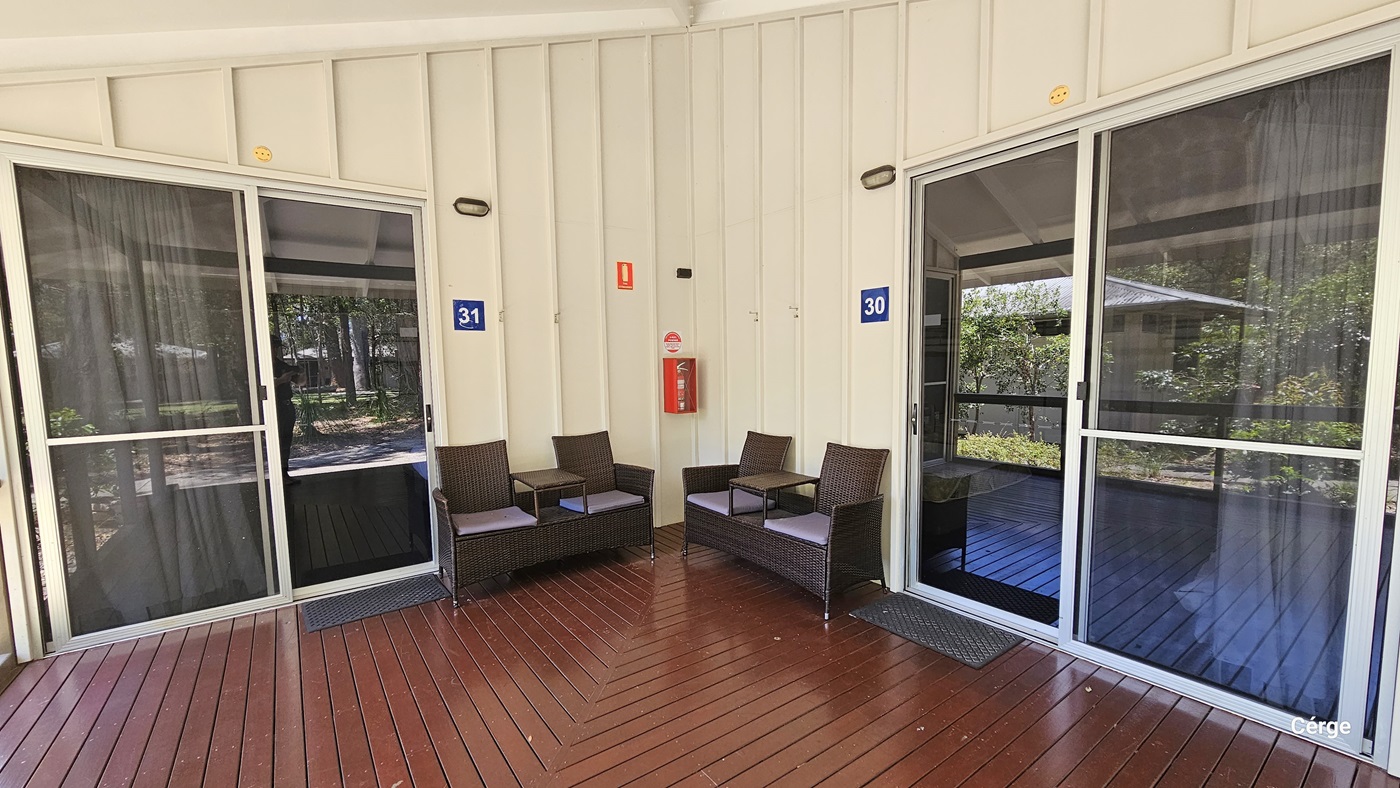 A low-rise building painted cream with corrugated roofing. The floor outside is made of polished wooden planks. There are two glass doors, each with a signage indicating Bangalow number. Rattan chairs are positioned in the corner, along with a fire extinguisher. Tall trees and shrubs surround the area.
