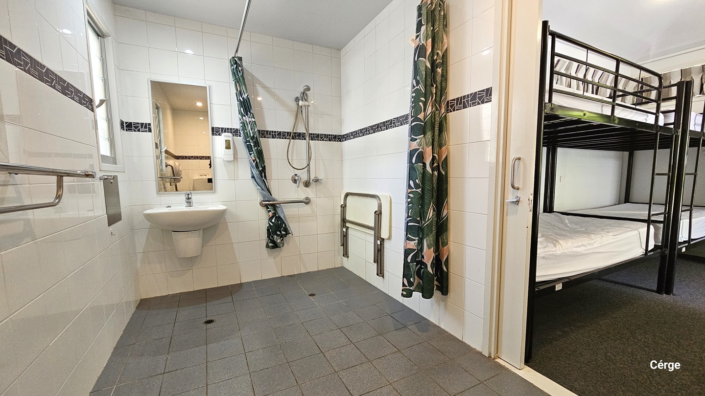 Inside the toilet. A hand grab, a wash basin, a mirror, and a soap dispenser are positioned on the left. On the right is a shower and green curtains. A foldable bench chair is mounted to the right wall. The wall has white tiles with a line of black prints as an accent. The ceiling is white, and the floor has grey tiles. To the right, double-deck beds are visible.
