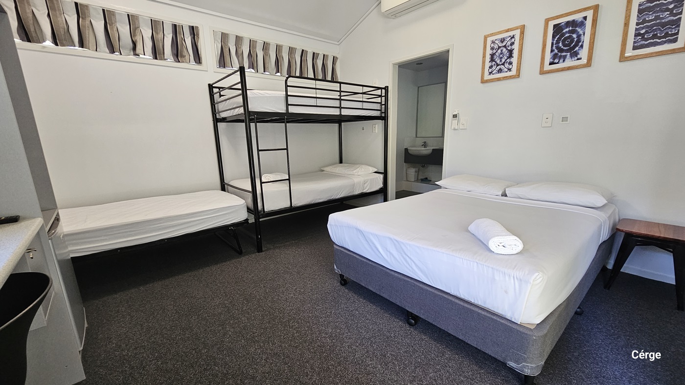 The ensuite room contains a single bed and a double-decker bed on the left, with a double bed adjacent. All beds are covered with white sheets. A small table is positioned on the right of the bed. Picture frames decorate the upper right wall. The floor has a dark grey carpet. The ceiling is white, and the left wall has short curtains. The toilet is positioned in the right corner.