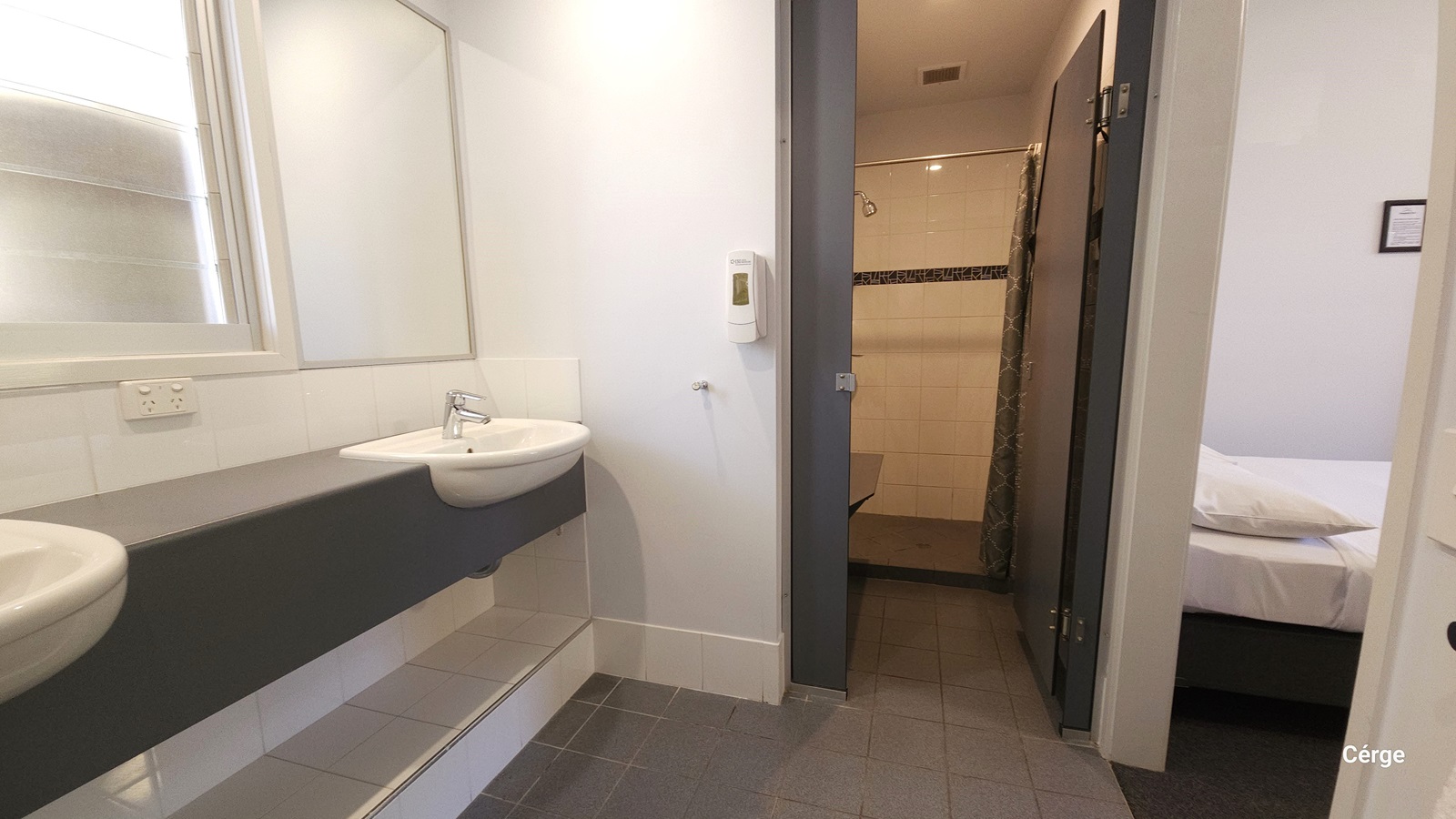 To the left are two wash basins and a mirror mounted to the left wall near a window. A soap dispenser is affixed to the middle wall. Adjacent is a door that leads to the toilet and shower area. To the right, a bed with white linen is visible.