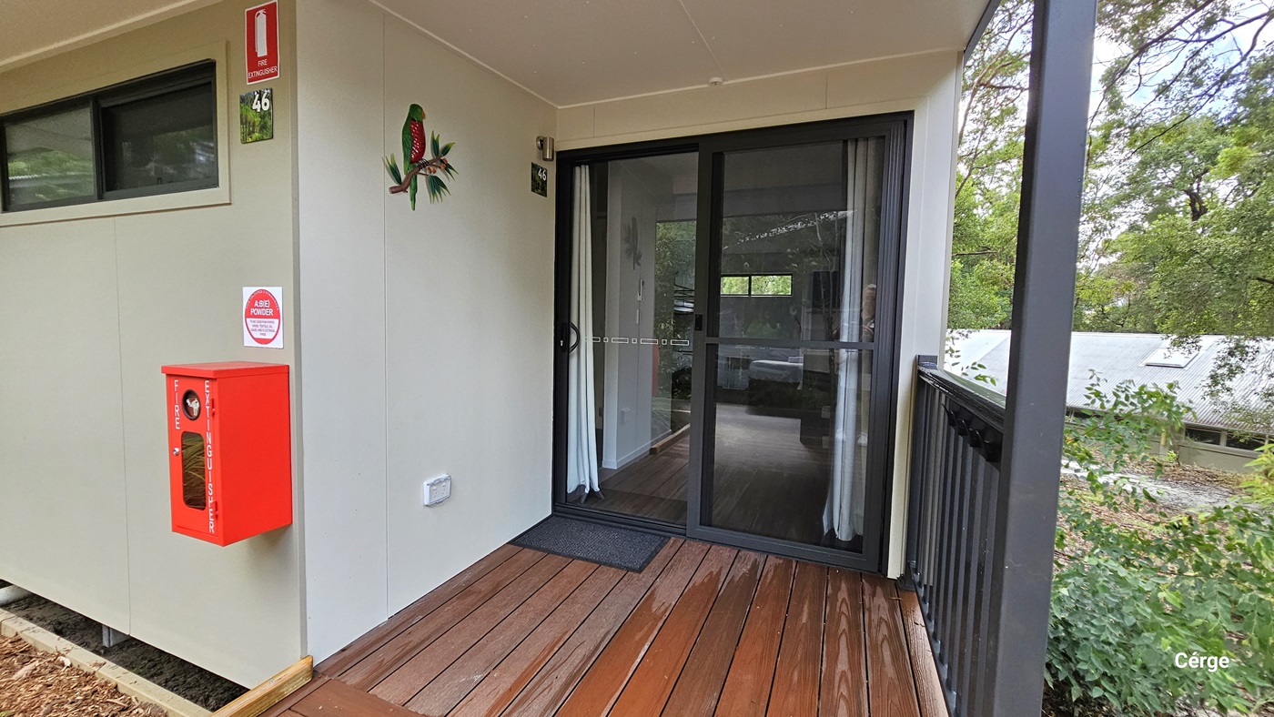 On the left, there is a beige wall with a window, a red fire extinguisher box, and a green parrot decoration. The middle features a glass sliding door with white curtains behind it, leading into an interior space. On the right, there is a black railing with greenery visible beyond it. The floor is made of wooden planks, and the ceiling is white and flat.