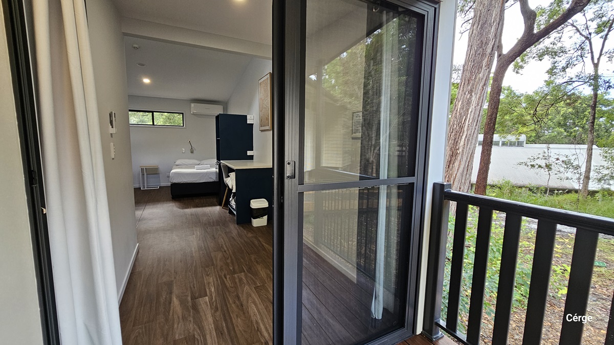 On the left, there is a narrow hallway with timber flooring leading to a bedroom. The bedroom contains a bed with white linens, a small bedside table, and a window above the bed. On the right, there is a glass sliding door opening to a balcony with a black railing. The balcony overlooks a garden area with trees and greenery. The ceiling is white and sloped, and the floor inside is timber.