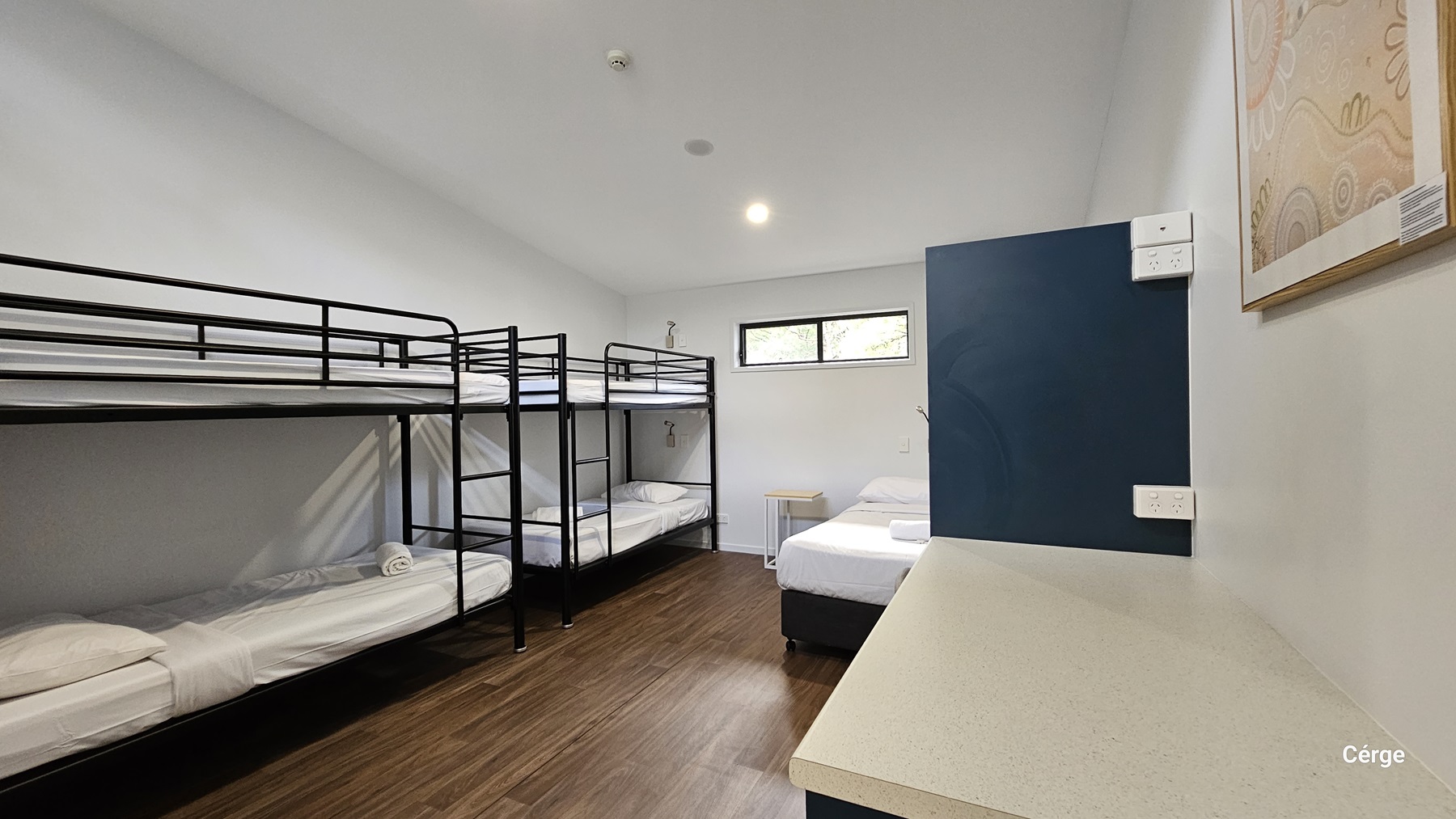 On the left, there are two sets of black metal bunk beds with white bedding. On the right, a blue door is partially open, revealing a single bed with white bedding. Above the bed, a small rectangular window lets in light. In the foreground on the right, a long white countertop extends along the wall. The floor is made of dark timber, and the ceiling is plain white with recessed lighting.