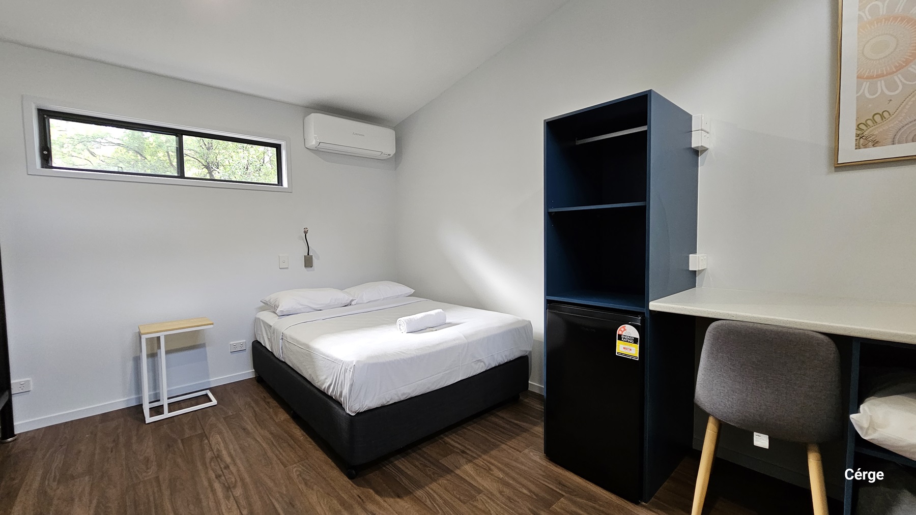 A room with a wooden floor and a slanted ceiling. On the left, there is a small window near the ceiling, an air conditioning unit below it, and a bed with white linens and a towel on top. Next to the bed is a small, minimalist side table. On the right, there is a dark blue shelving unit with a mini-fridge at the bottom, a desk with a chair, and a framed picture on the wall above the desk. The walls are painted white.