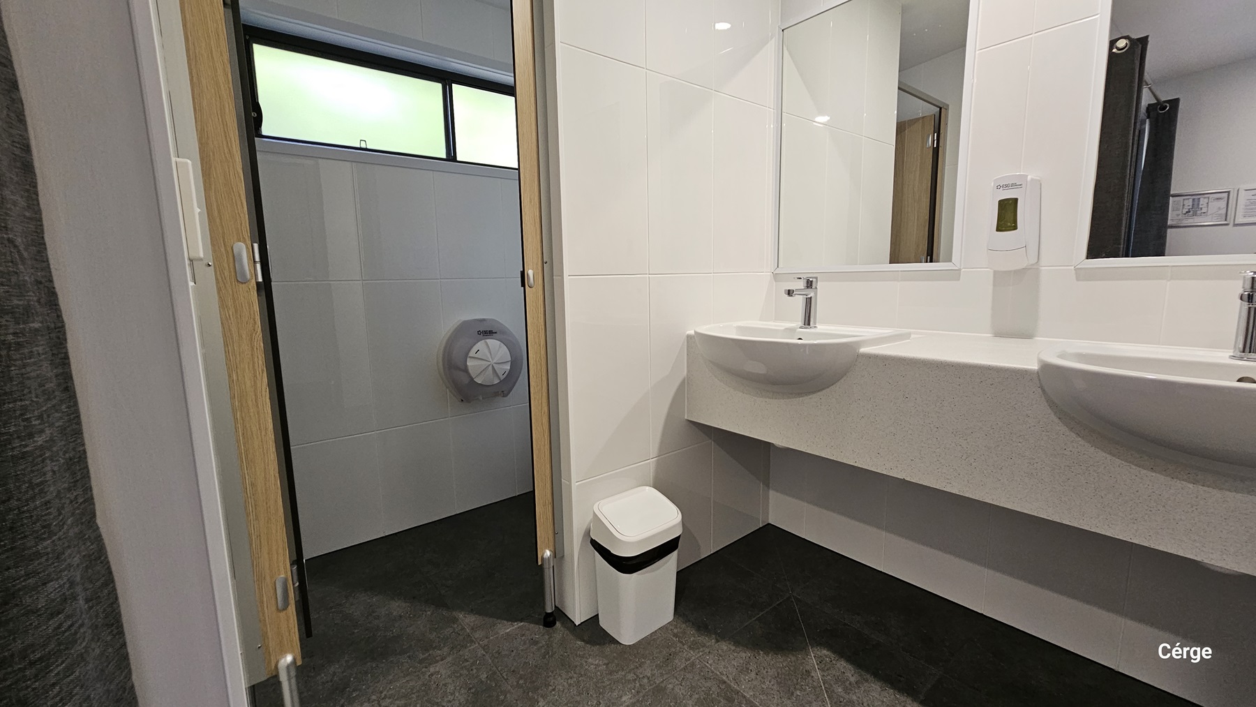 On the left, there is a partially open door revealing a toilet cubicle with a toilet paper dispenser mounted on the wall. The floor is dark grey tile. On the right, there are two white sinks with a soap dispenser mounted on the wall above them. A large mirror is positioned above the sinks. The background features white tiled walls. The ceiling is white and smooth. In the foreground, there is a small white bin placed under the sinks.