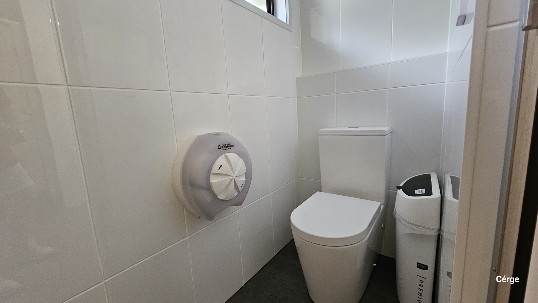 A small bathroom with white tiled walls. On the left, there is a large, round toilet paper dispenser mounted on the wall. In the middle, there is a white toilet with a rectangular cistern. To the right of the toilet, there is a white bin with a black lid. The floor is covered with dark grey tiles.
