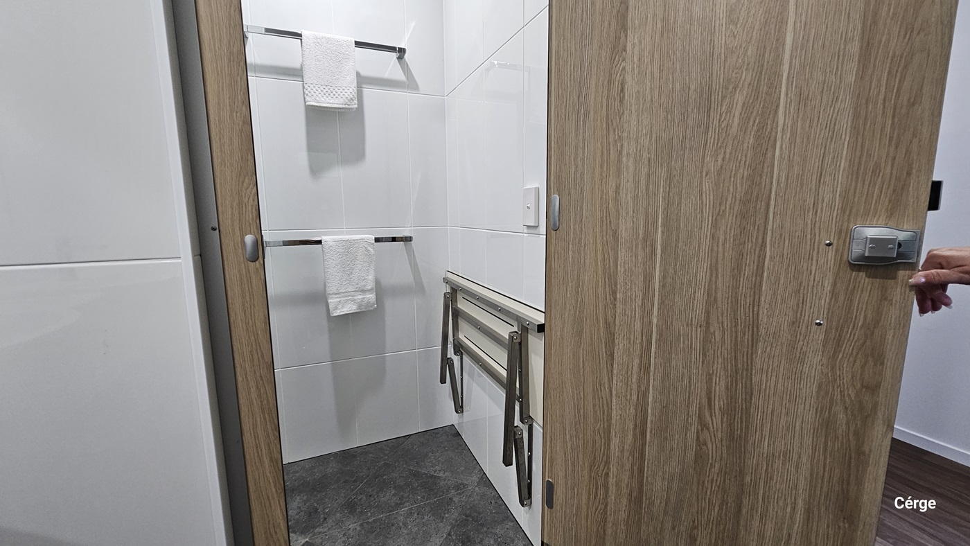 A small bathroom with white tiled walls. On the left, there is a white towel hanging on a metal rail. On the right, a wooden door is partially open. In the middle, there is a foldable shower bench attached to the wall. The floor is covered with dark grey tiles.