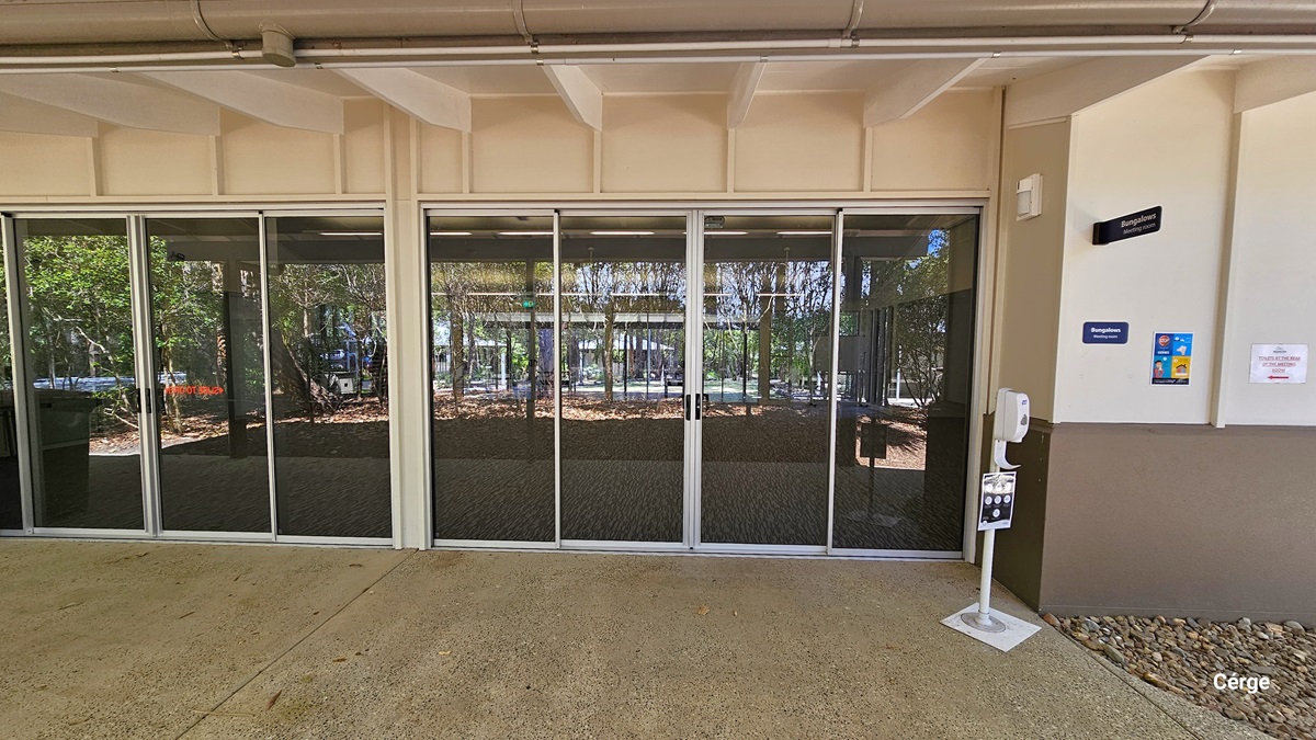 The building's front features automatic glass doors, with a hand sanitizer displayed to its right. Posters and signage are affixed to the white wall. The ground is covered with gravel on the right and concrete on the left.