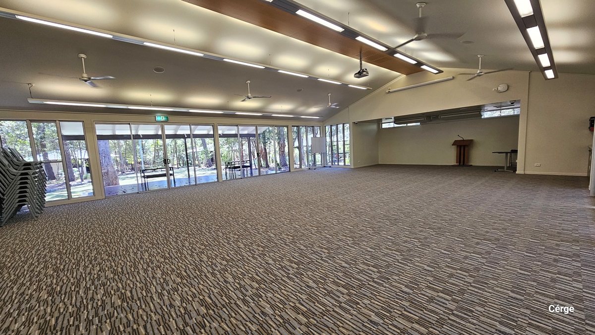 A spacious, empty hall with printed grey carpet. The pitched ceiling features rows of fluorescent lights and ceiling fans. A brown wooden table stands at the far end. Glass walls and glass doors cover the left side. Plastic chairs are stacked on the left.