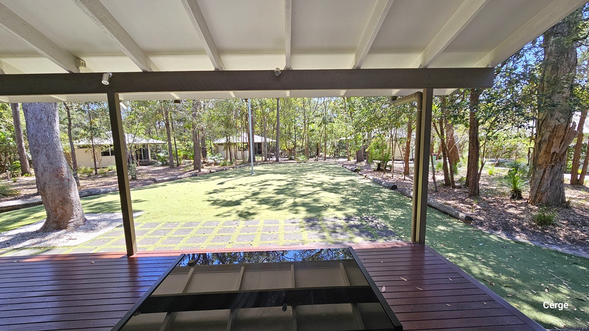 A spacious area covered with green artificial turf bordered by logs. A tall steel post is positioned in the centre. In the middle stands a low-rise cream building made of concrete and glass. Tall trees surround the area.