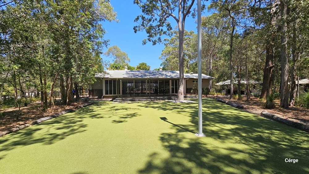 A low set, cream building set amongst tall trees with a large green, grassed area in the foreground.