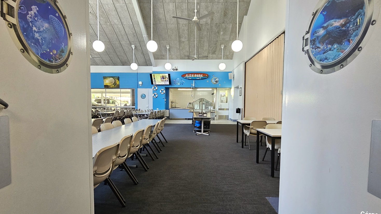 A spacious dining hall with nautical-themed decor. The room features long rows of white tables with attached bench-style seating. The floor has a dark grey carpet, and the ceiling has a grey finish. Pendant lights and ceiling fans hang from above. On the walls, there are two circular paintings resembling portholes. Additionally, there are framed pictures and a large wall clock above a serving counter at the far end of the room.