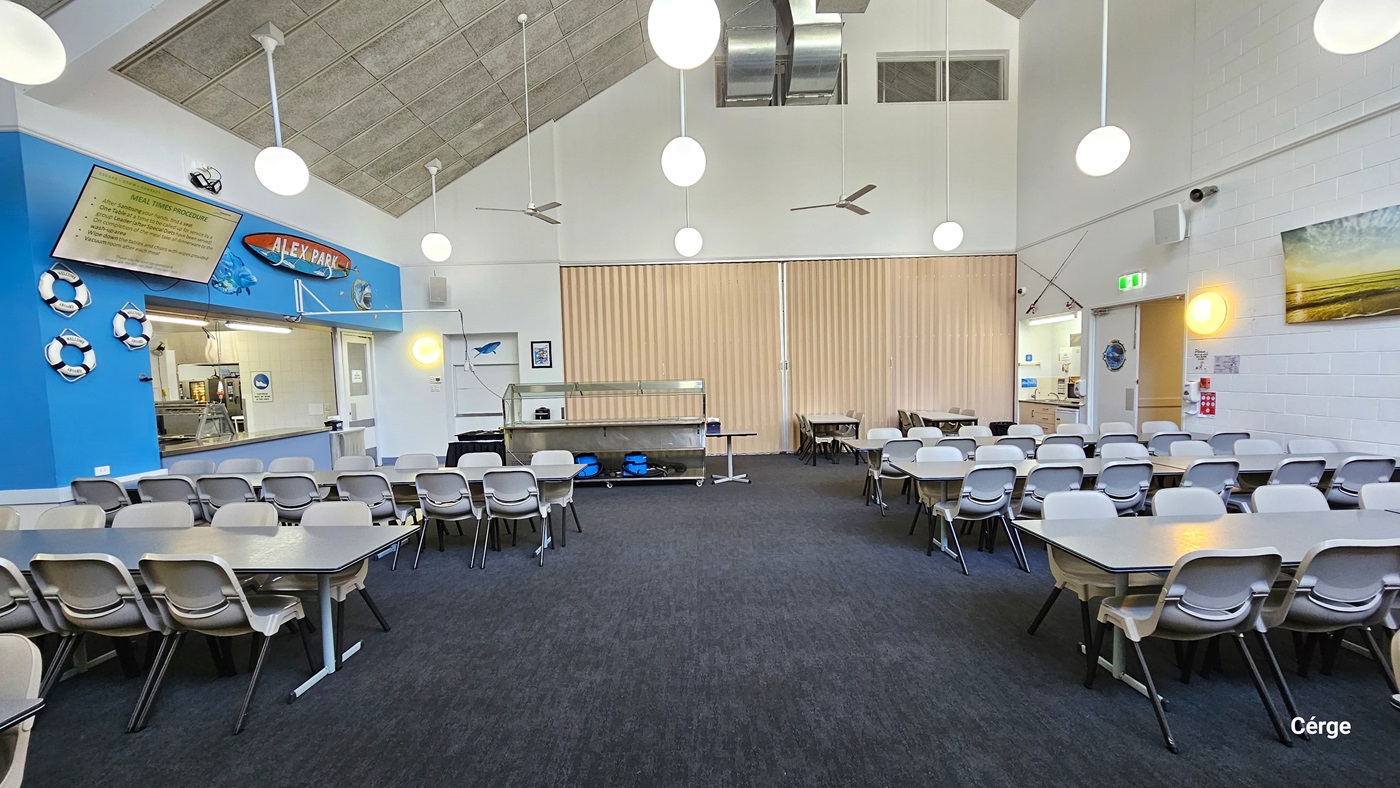 A spacious dining hall with nautical-themed decor. The room features rows of white tables with attached bench-style seating. The floor has dark grey carpet, and the pitched ceiling has a grey finish. Pendant lights and ceiling fans hang from above. On the left, there are framed pictures, a flatscreen TV, and framed posters positioned on top of the serving counter. A retractable wall separator is positioned in the middle.