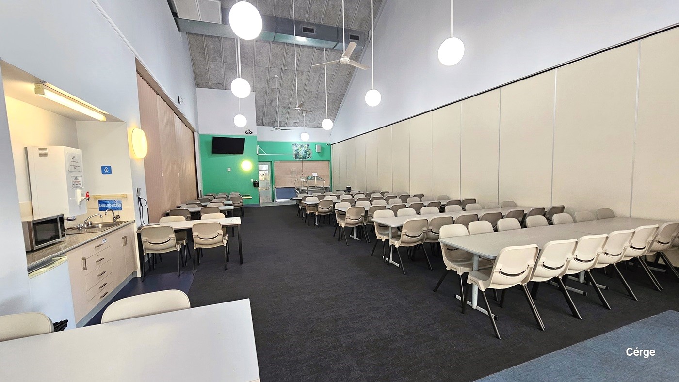 The dining room features rows of white tables with attached bench-style seating. The floor has dark grey carpet, and the pitched ceiling has a grey finish. Pendant lights and ceiling fans hang from above. On the left, there is a counter table holding a microwave oven, a wash basin, and a water dispenser. A retractable wall separator is positioned on the right.