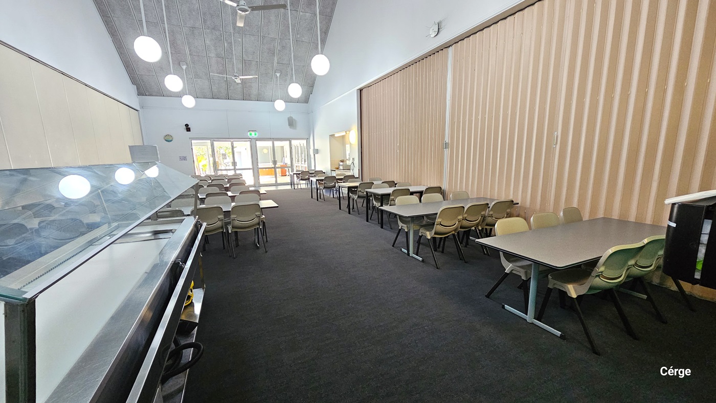 The dining room features rows of white tables with attached bench-style seating. The floor has a dark grey carpet, and the pitched ceiling has a grey finish. Pendant lights and ceiling fans hang from above. On the left, there is a food display rack. A cream retractable wall separator is positioned on the right. A glass door is positioned at the far end.