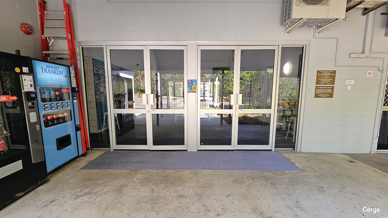 A pair of double glass doors set in a light grey wall within a building, flanked by red fire extinguisher equipment to the left and a vending machine for cold drinks. An orange ladder is placed against the wall. A striped mat lies in front of the doors. Signs and notices are visible on the wall next to the door, and the interior behind the glass doors appears to be a foyer or entrance hall with seating.