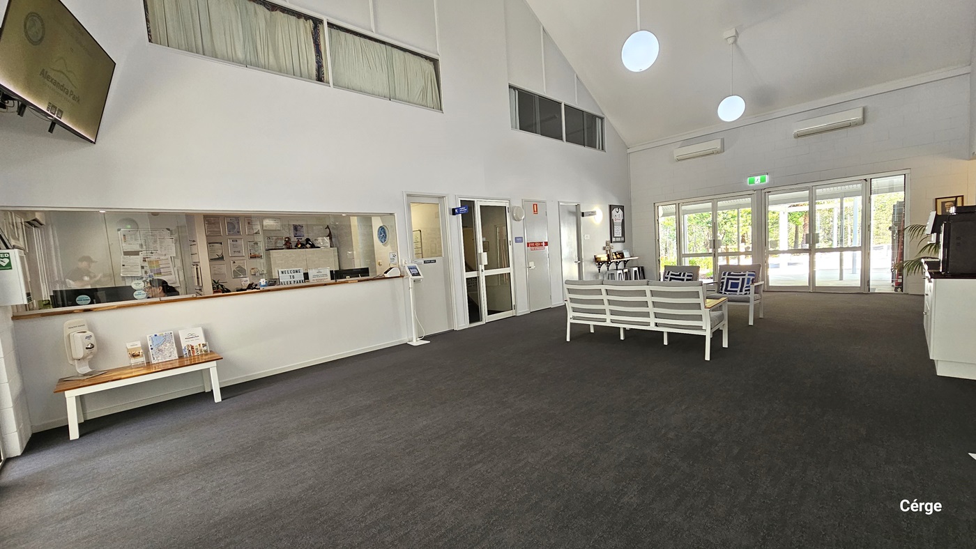 A spacious interior of a building with grey carpet flooring and white walls. There's a reception desk to the left with various pamphlets displayed on a wooden table. Above it is a flatscreen TV. A seating area with cushioned grey chairs and a table is positioned in the centre. Pendant lights hang from the ceiling. Behind is a set of automatic glass doors with an exit sign above.