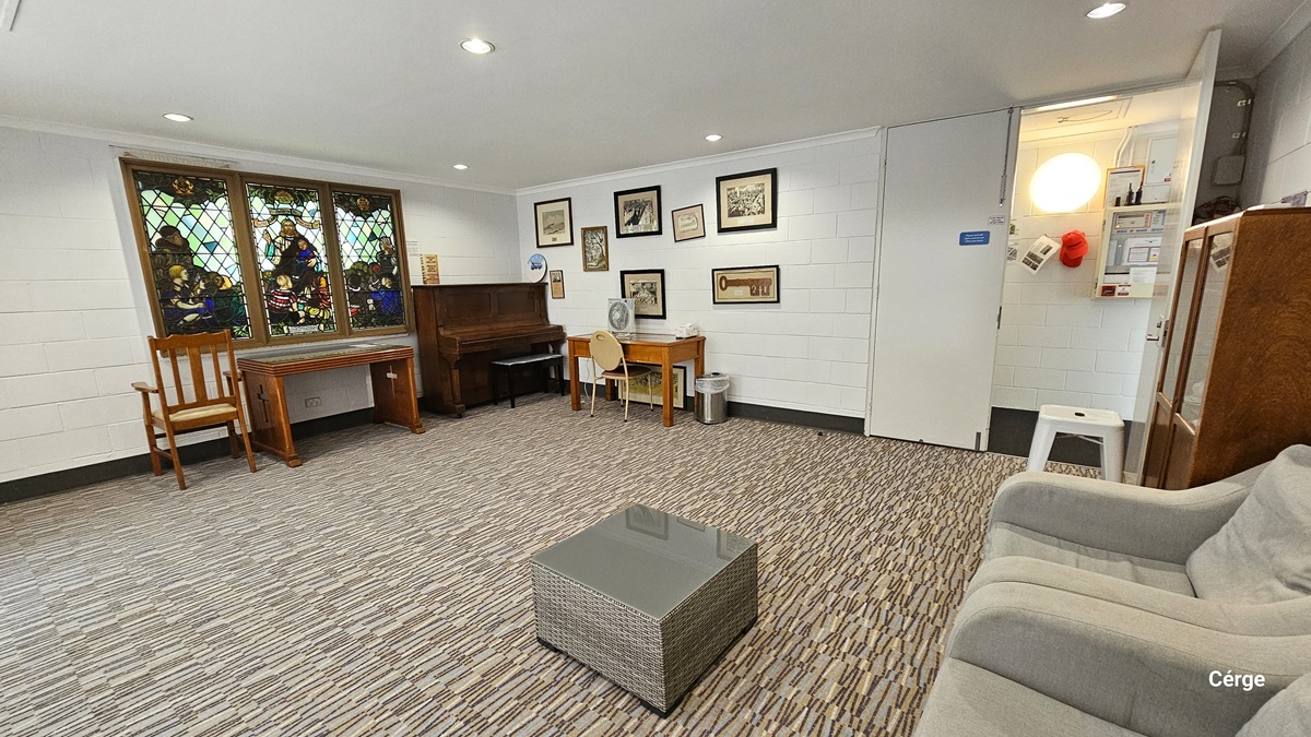 In the corner is a wooden piano. Beside it is a stained glass window with a painting, along with wooden tables and chairs. In the middle is a white wall adorned with framed pictures and some memorabilia. Another wooden table is placed against it. On the right, there is a gray sofa along with a small table and a wooden display rack. The ceiling is all white with LED lights, and the floor has printed carpets.